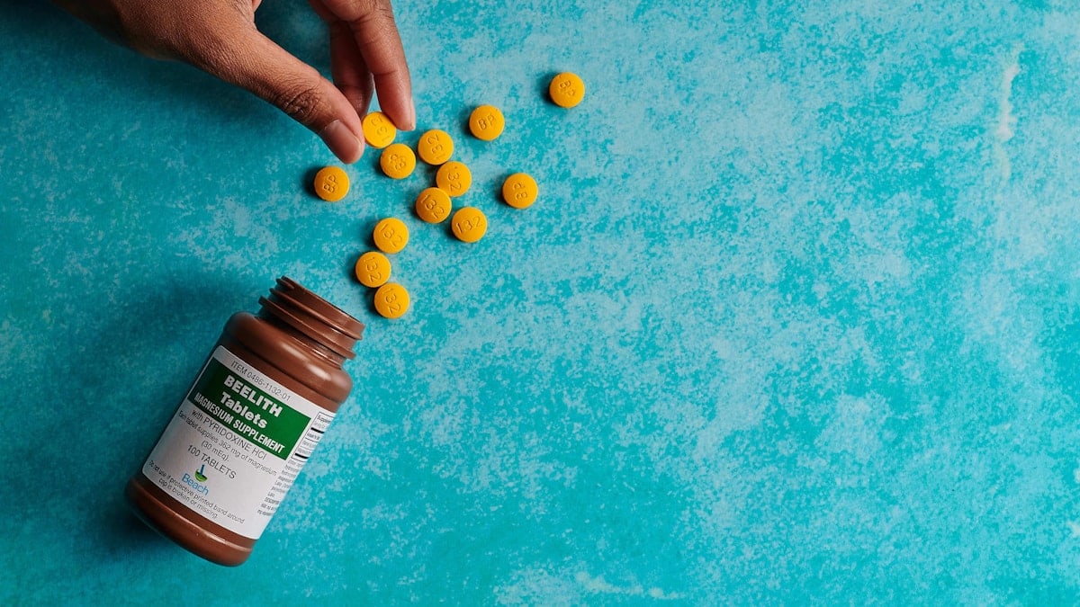 hand holding a yellow pill of medicine next the the bottle and a pile of pills